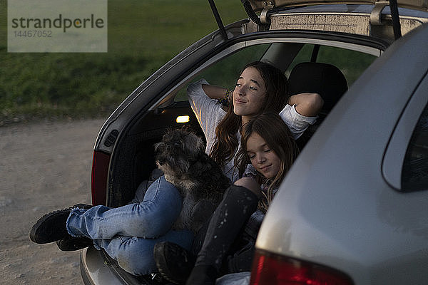 Zwei Schwestern im geöffneten Kofferraum eines geparkten Autos beim Sonnenuntergang