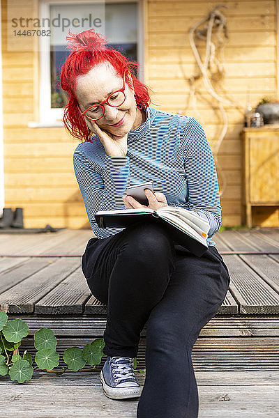 Porträt einer lächelnden älteren Frau mit rot gefärbten Haaren  die mit ihrem Handy auf der Terrasse vor ihrem Haus sitzt