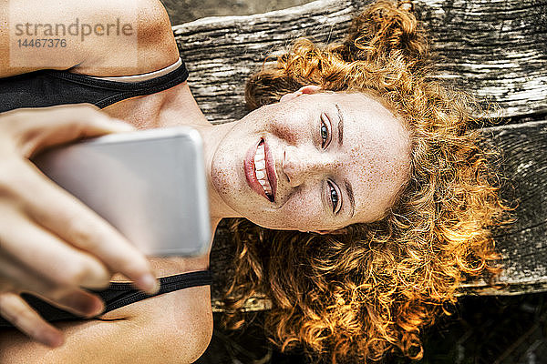 Porträt einer rothaarigen jungen Frau  die mit ihrem Handy auf einer Bank liegt