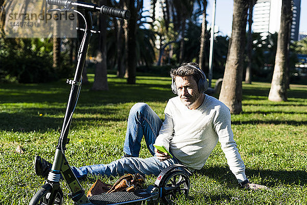 zufälliger Geschäftsmann  der auf einem Rasen in einem Park sitzt und ein Smartphone und Kopfhörer benutzt