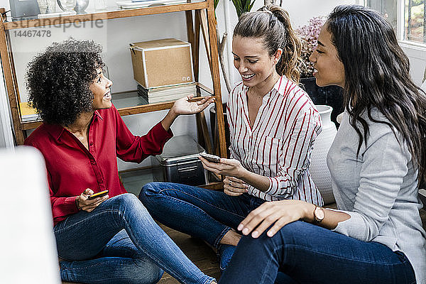 Drei glückliche Frauen sitzen zu Hause mit Mobiltelefonen auf dem Boden