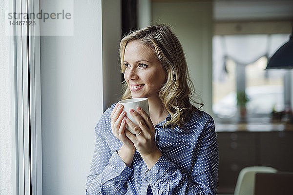 Lächelnde Frau hält zu Hause eine Tasse Kaffee am Fenster