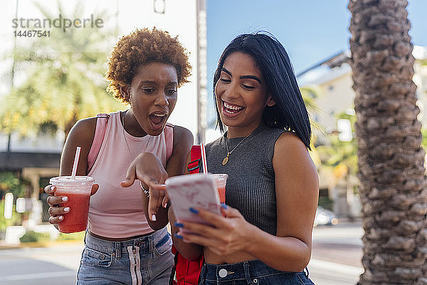 USA  Florida  Miami Beach  zwei glückliche Freundinnen mit Handy und Erfrischungsgetränk in der Stadt