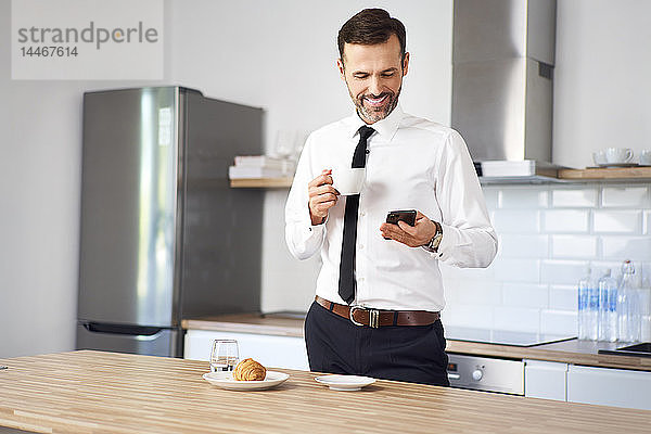Mann  der vor der Arbeit in der Küche steht  Kaffee trinkt und auf sein Telefon schaut