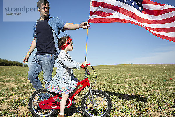 Mann und Tochter mit Fahrrad und amerikanischer Flagge auf Feld in entlegener Landschaft