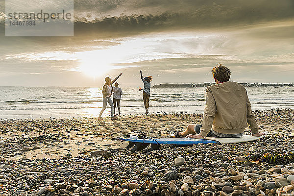 Junger Mann auf dem Surfbrett beobachtet Freunde beim Seifenblasenmachen am Strand bei Sonnenuntergang