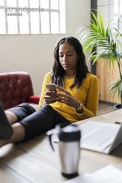 Junge Geschäftsfrau sitzt mit den Füßen auf dem Schreibtisch und benutzt ein Mobiltelefon