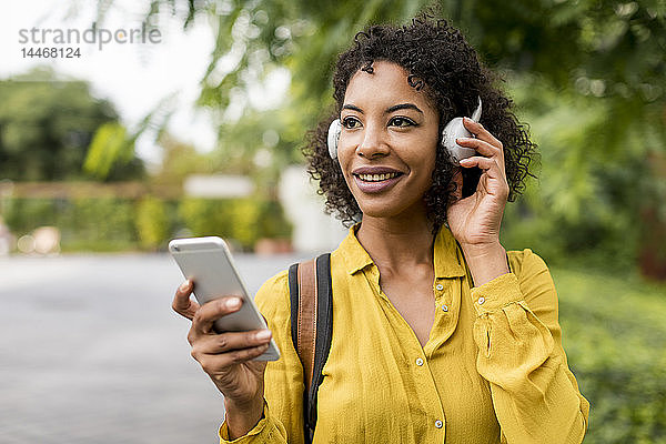 Porträt einer lächelnden Frau  die im Freien mit Kopfhörern und Smartphone Musik hört