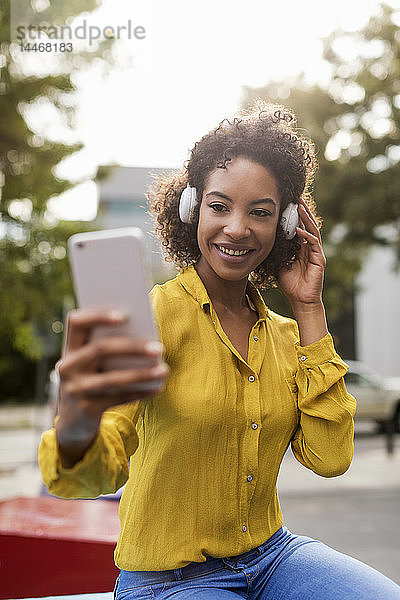 Porträt einer lächelnden Frau  die mit Kopfhörern Musik hört und sich mit einem Smartphone selbstständig macht