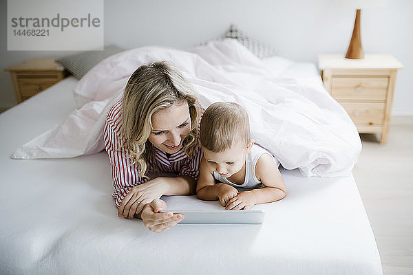 Lächelnde Mutter und Kleinkind-Sohn liegen zu Hause im Bett und benutzen Tabletten