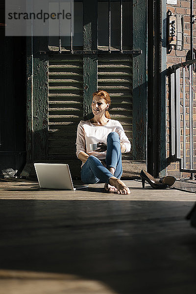 Lächelnde Geschäftsfrau sitzt auf dem Boden mit Tasse Kaffee und Laptop