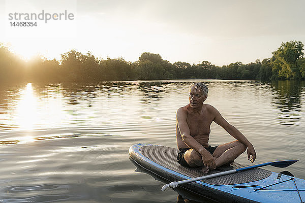 Älterer Mann sitzt bei Sonnenuntergang im SUP-Vorstand