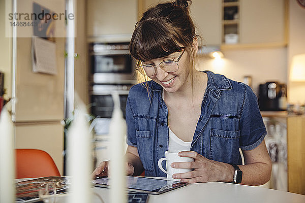 Lächelnde Frau sitzt mit einer Tasse Kaffee am Tisch in der Küche und benutzt Tabletten