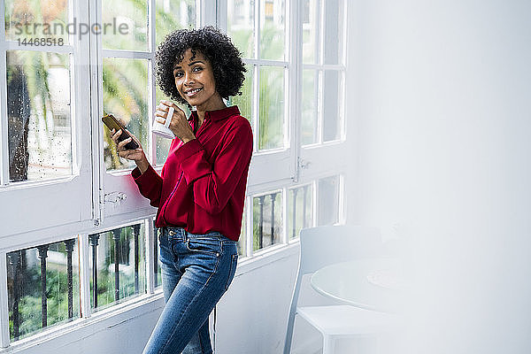 Porträt einer lächelnden Frau mit Tasse Kaffee und Handy  die zu Hause am Fenster steht