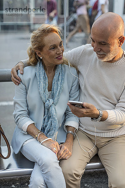 Spanien  Barcelona  ein älteres Ehepaar sitzt an einer Straßenbahnhaltestelle in der Stadt und teilt sich ein Smartphone mit Ohrstöpseln
