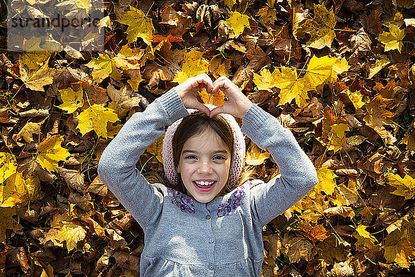 Porträt eines fröhlichen kleinen Mädchens  das auf Herbstlaub liegt und mit den Händen das Herz formt