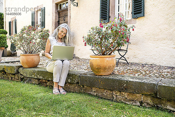 Frau mit langen grauen Haaren sitzt mit Laptop auf der Terrasse eines Landhauses