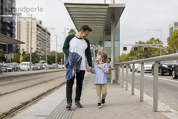 Lächelnder Vater und Sohn gehen Hand in Hand an der Straßenbahnhaltestelle in der Stadt