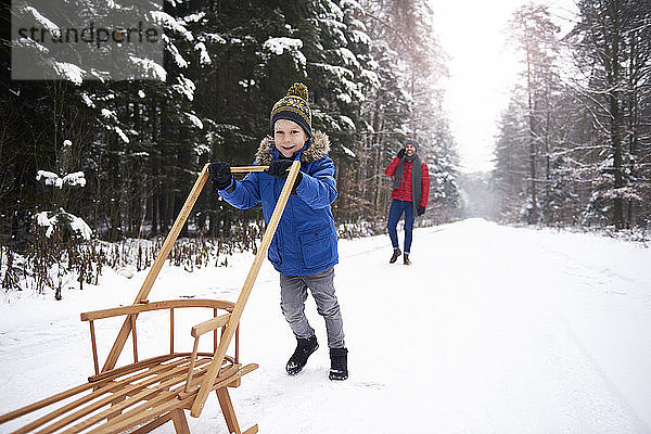 Kleiner Junge schiebt Schlitten im Winterwald  während sein Vater ihn aus dem Hintergrund beobachtet