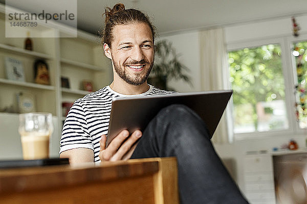 Junger Mann mit einem Brötchen zu Hause sitzend  mit digitalem Tablet