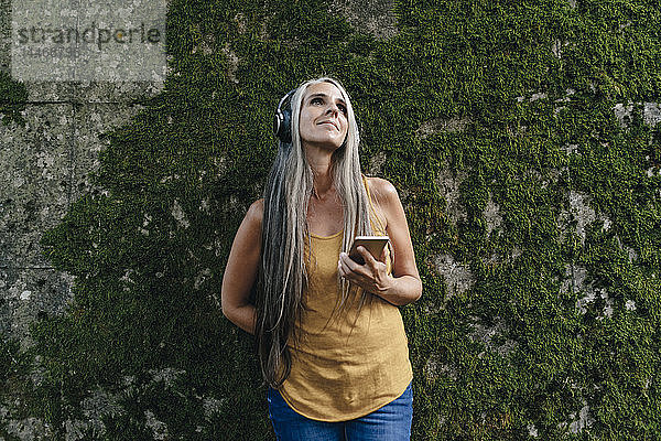Frau mit Handy steht vor der Wand und hört Musik mit Kopfhörern