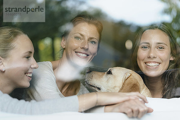 Porträt einer glücklichen Mutter mit zwei Teenager-Mädchen und Hund hinter einer Fensterscheibe