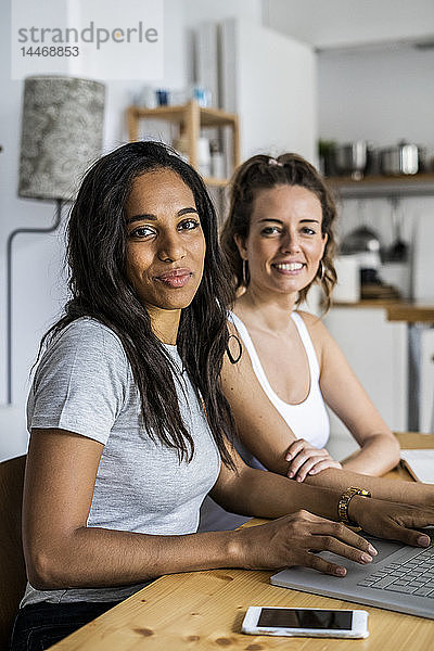 Porträt von zwei lächelnden Frauen  die mit Laptop zu Hause am Tisch sitzen