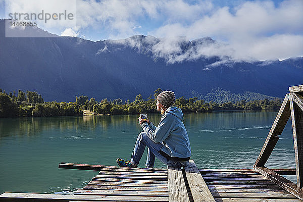 Chile  Chaiten  Lago Rosselot  Frau sitzt auf dem Steg und hält einen Becher