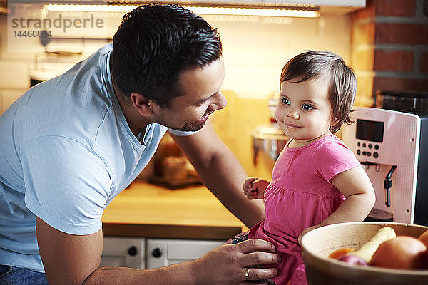 Lächelnder Vater sieht Baby-Mädchen an  das zu Hause in der Küche auf dem Tresen sitzt