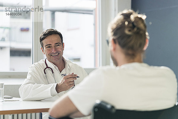 Lächelnder Arzt im Gespräch mit Patient im Rollstuhl in der medizinischen Praxis