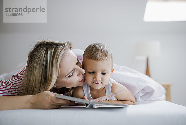 Mutter küsst Kleinkind Sohn  der zu Hause im Bett liegt und ein Buch liest