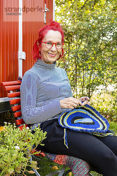 Porträt einer lächelnden älteren Frau mit rot gefärbten Haaren  die im Garten vor einem roten Wohnwagen sitzt und häkelt