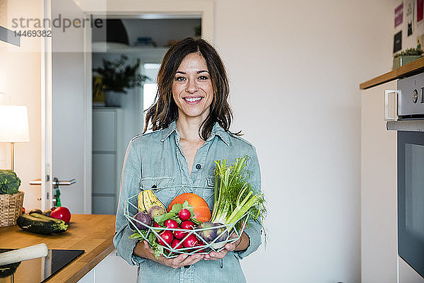 Frau steht in der Küche und hält einen Korb mit frischem Obst und Gemüse