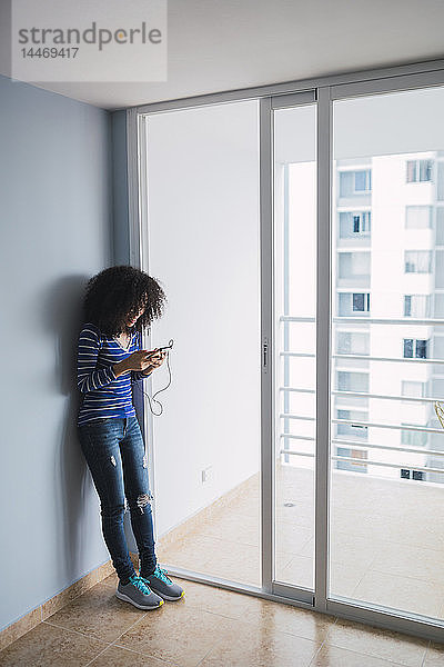Lächelnde junge Frau steht an der geöffneten Balkontür ihrer neuen Wohnung und benutzt ein Smartphone