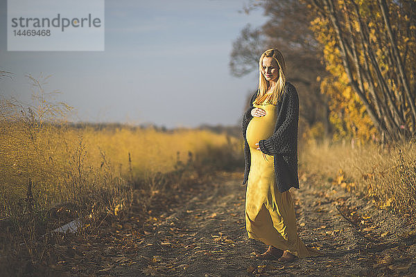 Lächelnde schwangere Frau steht im Herbst auf einem Waldweg neben einem Spargelfeld