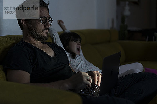 Vater mit Tochter sitzt nachts mit Laptop auf der Couch