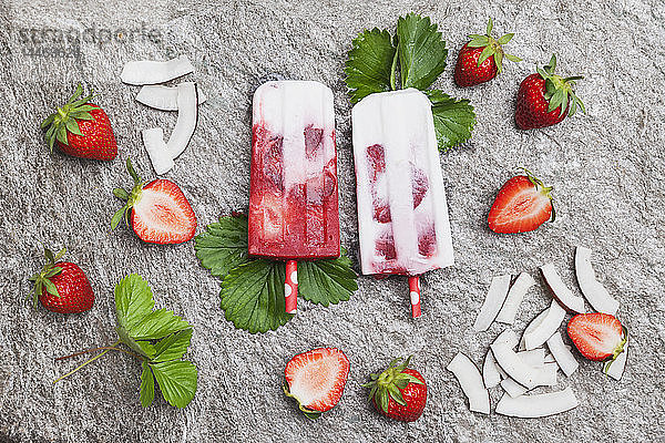 Hausgemachte Erdbeer-Kokos-Eis-Lollies mit frischen Erdbeeren und Kokosnussscheiben auf Granit