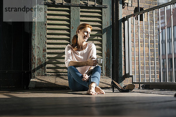 Lächelnde Geschäftsfrau sitzt barfuss mit einer Tasse Kaffee auf dem Boden