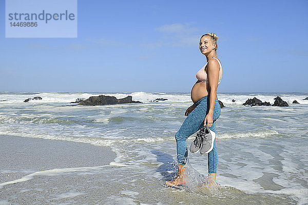 Lächelnde schwangere Frau am Strand beim Waten im Meer