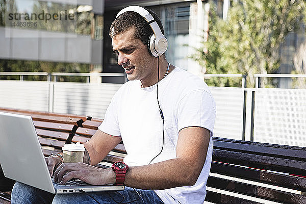 Junger Mann sitzt auf einer Bank  trägt Kopfhörer und benutzt einen Laptop