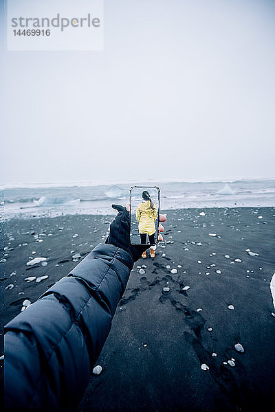 3D-Montage eines Mannes  der mit einem Smartphone ein Bild der isländischen Landschaft aufnimmt  und einer Frau in gelbem Regenmantel