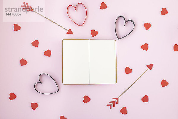 Valentinstagssymbole und aufgeklapptes Notizbuch auf rosa Hintergrund