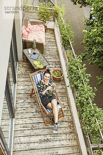 Junge Frau sitzt im Liegestuhl und entspannt auf ihrem Balkon