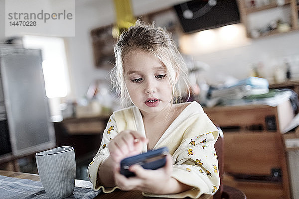 Porträt eines kleinen Mädchens  das mit einem Smartphone am Frühstückstisch in der Küche sitzt