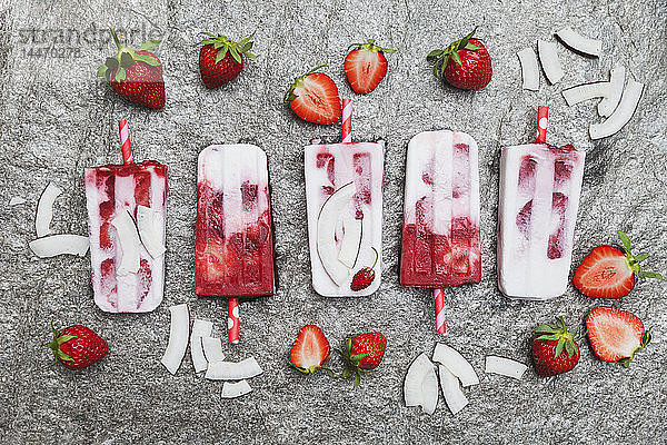 Hausgemachte Erdbeer-Kokos-Eis-Lollies mit frischen Erdbeeren und Kokosnussscheiben auf Granit
