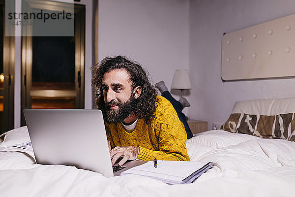 Lächelnder junger Mann liegt zu Hause im Bett und benutzt einen Laptop