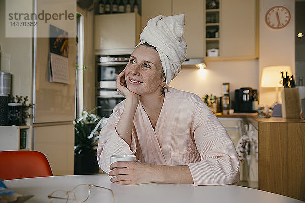 Porträt einer lächelnden Frau mit Handtuchturban  die mit einer Tasse Kaffee bei Tisch in der Küche sitzt