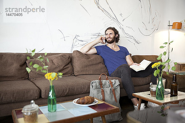 Porträt eines jungen Mannes am Telefon  der in einem Café mit Buch auf der Couch sitzt