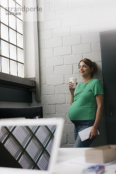 Lächelnde schwangere Frau mit Einwegbecher  die im Büro an einer Wand lehnt