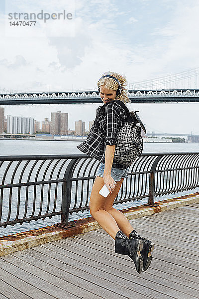 USA  New York City  Brooklyn  fröhliche junge Frau mit Kopfhörern und Handy am Wasser springend
